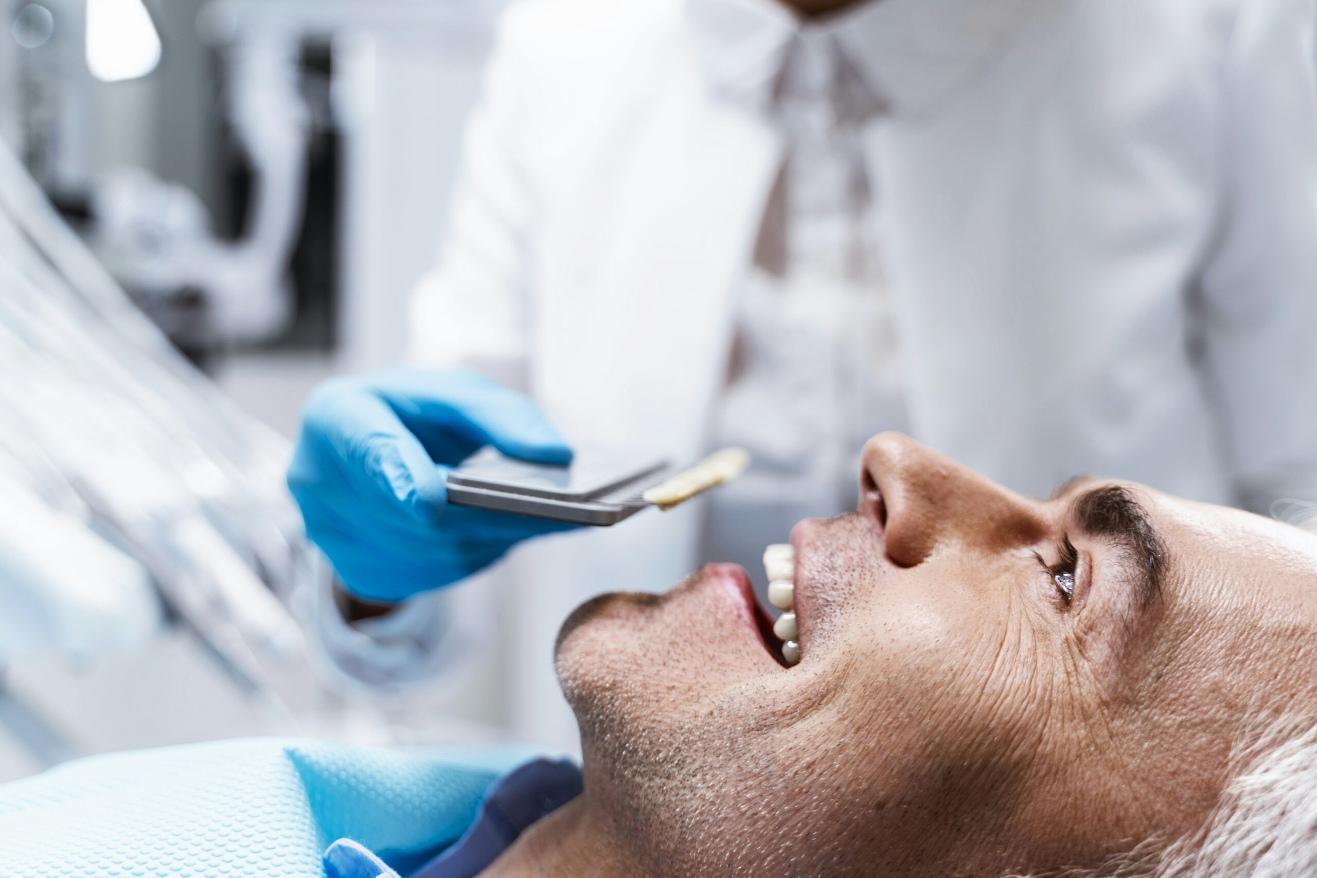 A dentist wearing blue gloves holds a shade guide near the smiling mouth of a male patient reclining in a dental chair, assessing his tooth color for matching or whitening.