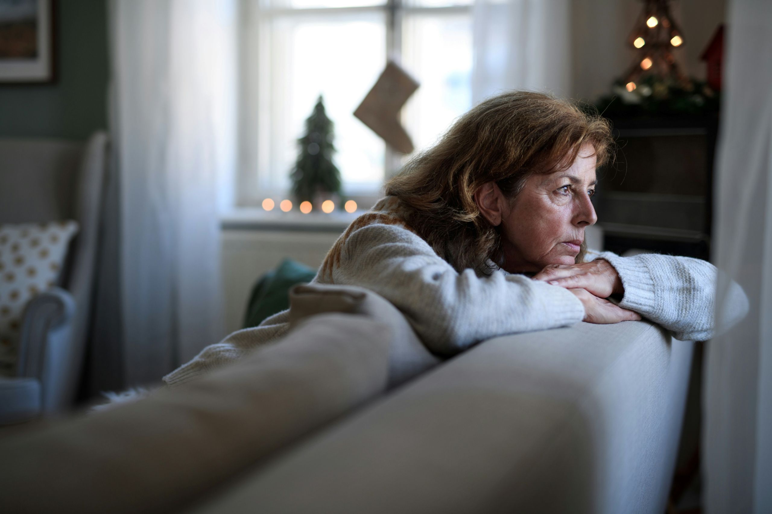 A woman leans on the back of a sofa, gazing thoughtfully out a window with her arms crossed. Soft daylight and holiday decorations, including a lit Christmas tree, create a cozy mood—perfect for reflecting on new resolutions like dental health tips.
