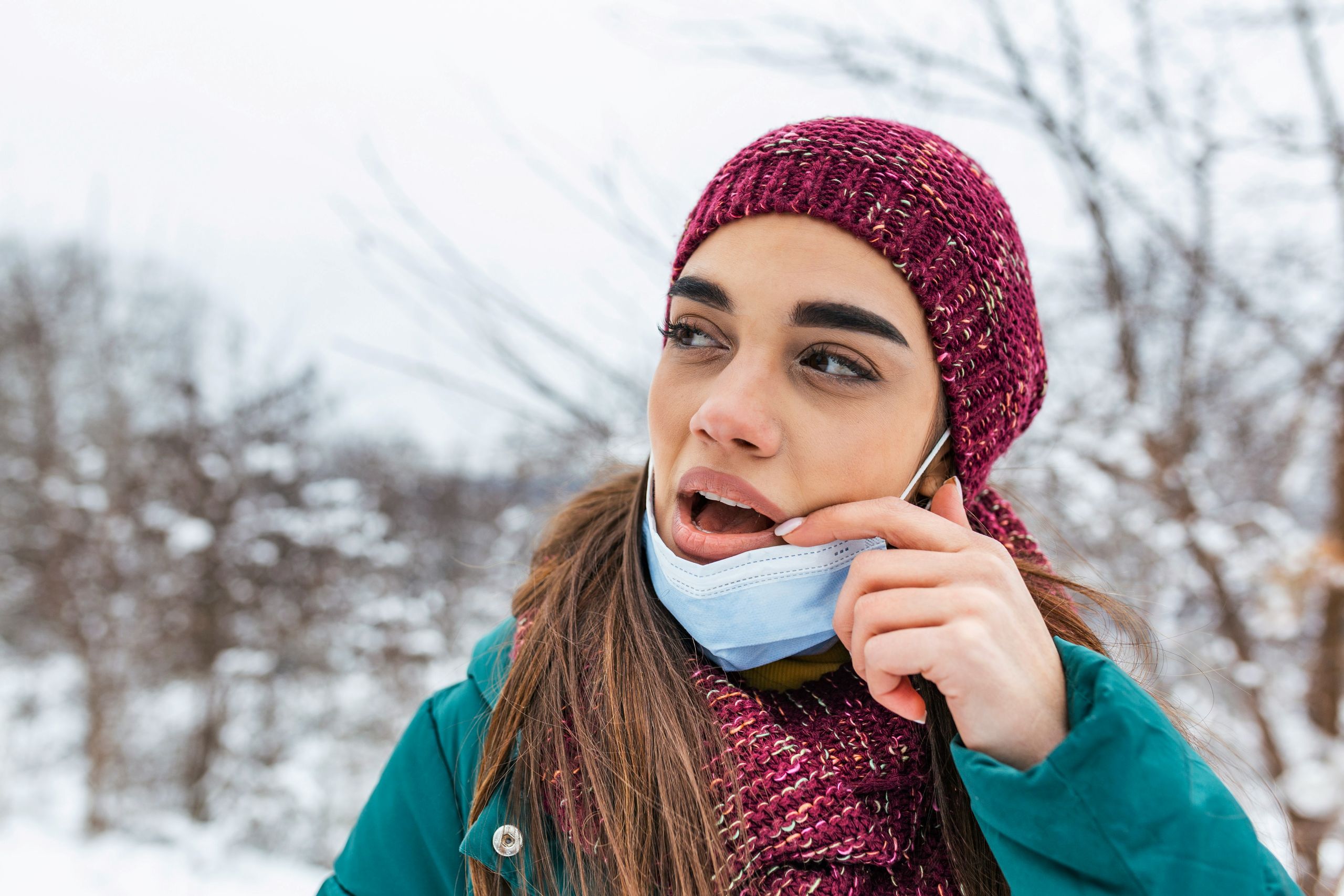 A young woman outdoors in winter, showing winter sensitivity, wears a red knit hat, scarf, and teal coat as she pulls down her blue face mask while looking to the side. Snowy trees are visible in the blurred background.