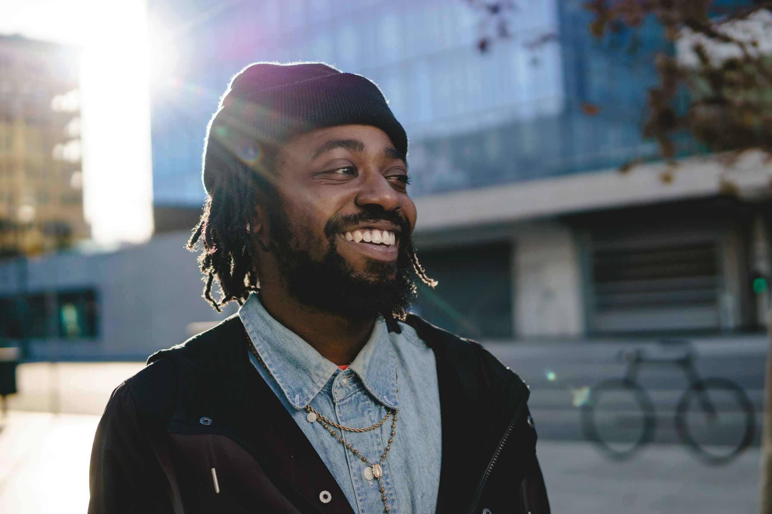 A smiling man, who could be a dentist, wears a black beanie, dark jacket, and layered necklaces as he stands outdoors in sunlight, modern glass buildings and a bicycle softly blurred in the background.