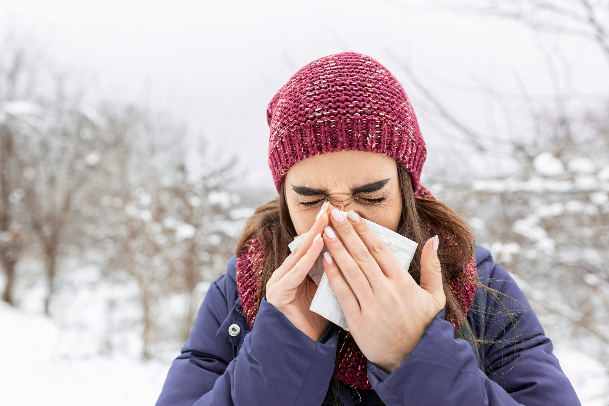 A person wearing a red knit hat and scarf blows their nose with a tissue while standing outdoors in a snowy, winter landscape, possibly experiencing the discomfort of winter toothaches.