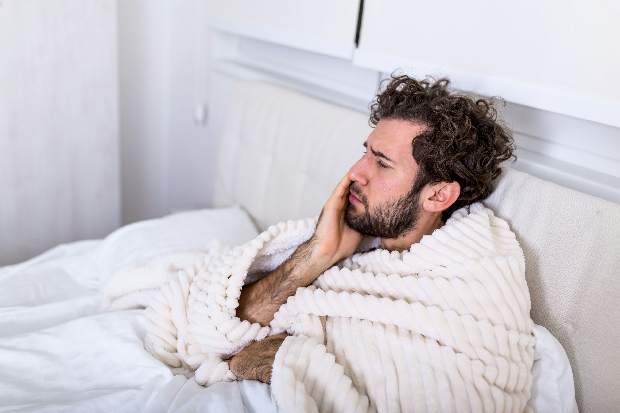 A man with curly hair sits up in bed, wrapped in a white blanket, holding his cheek with a pained expression, clearly suffering from tooth nerve pain.