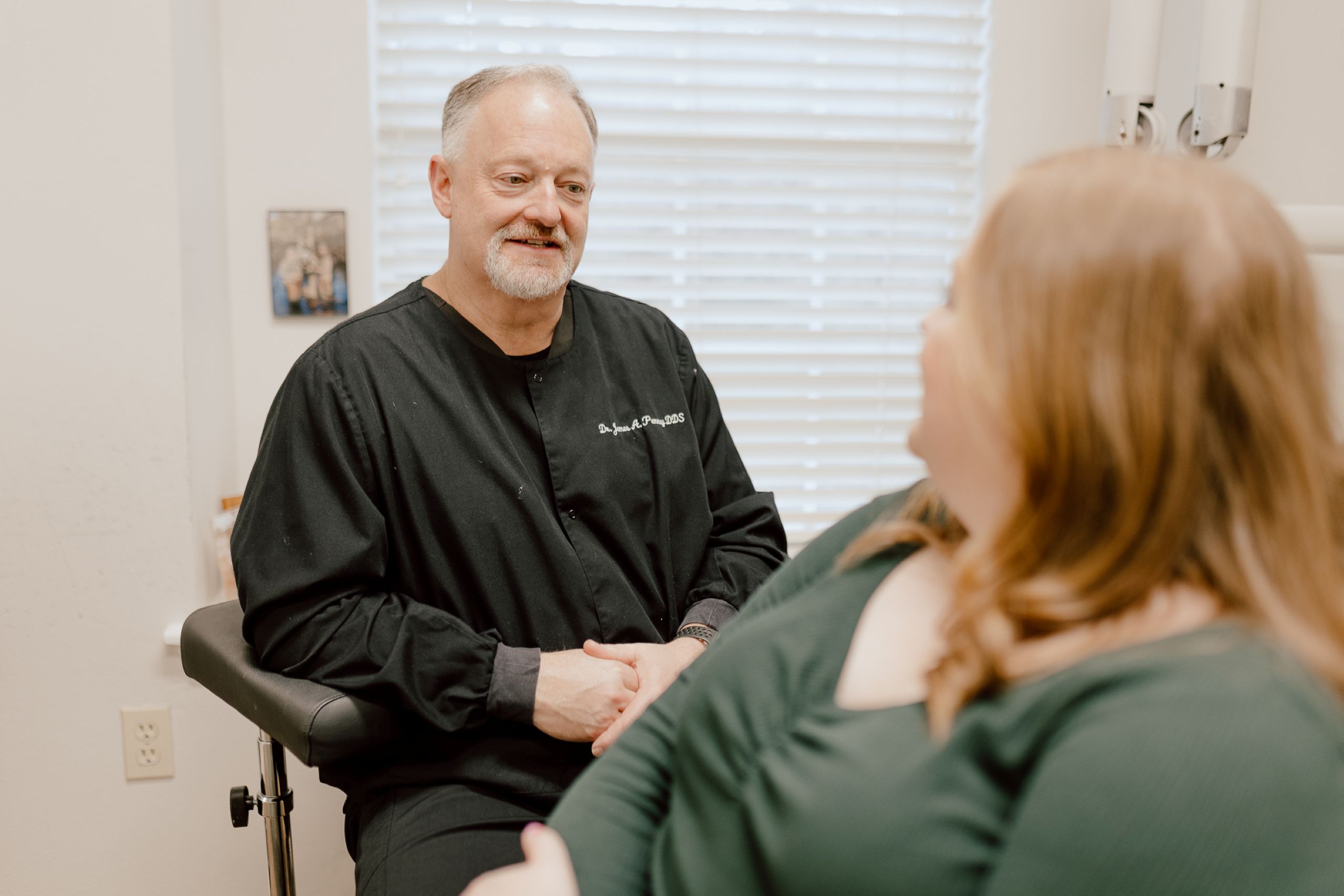 A male dentist in a black coat sits and talks with a female patient in a clinic room. The dentist is smiling, and they are positioned near a window with blinds. The patient is facing the dentist.
