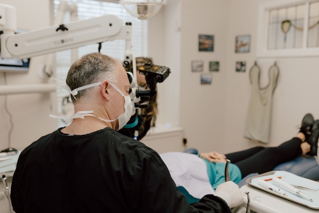 A dentist wearing a face mask and magnifying glasses examines a patient who is lying back in a dental chair in a bright, clean clinic room.