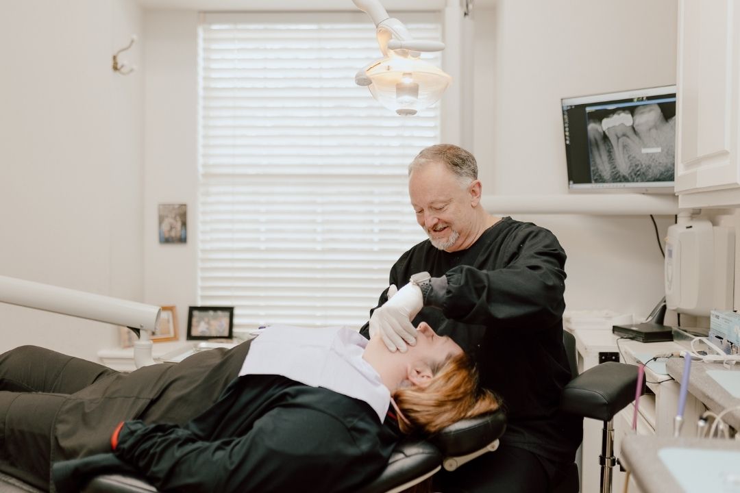 A dentist in black scrubs examines a patient lying in a dental chair. The dentist smiles while working. An X-ray image is displayed on a monitor in the background, and there is bright natural light from a window.