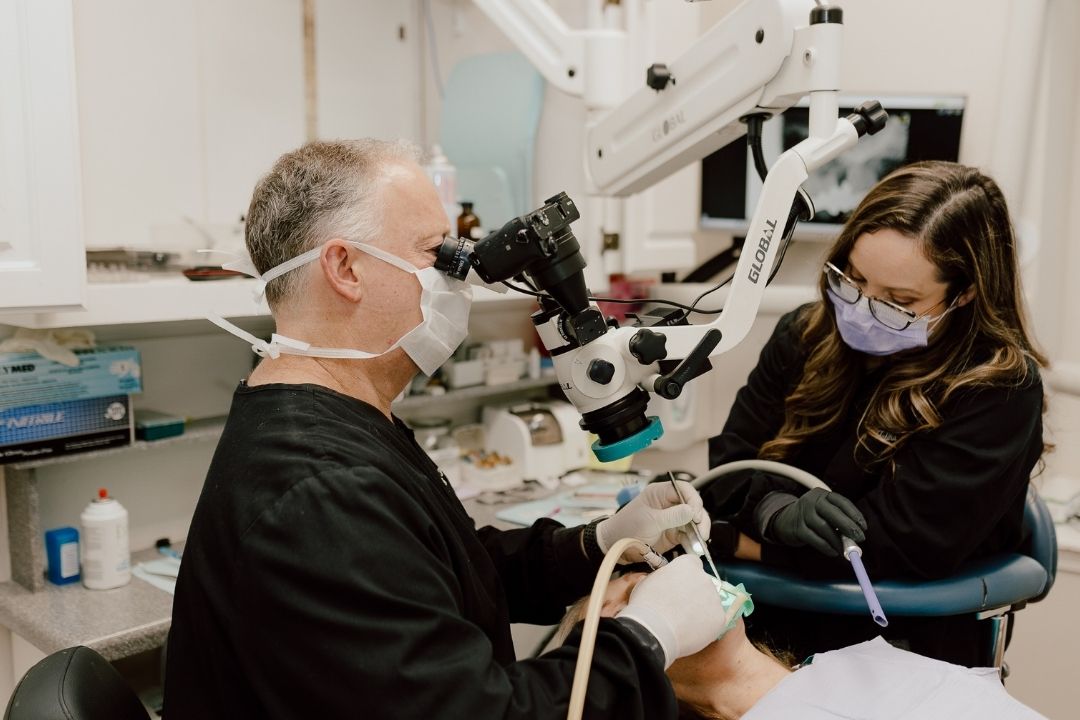 A dentist wearing a mask uses a microscope while treating a patient, assisted by a dental hygienist in a mask and gloves. They are working in a modern dental clinic.