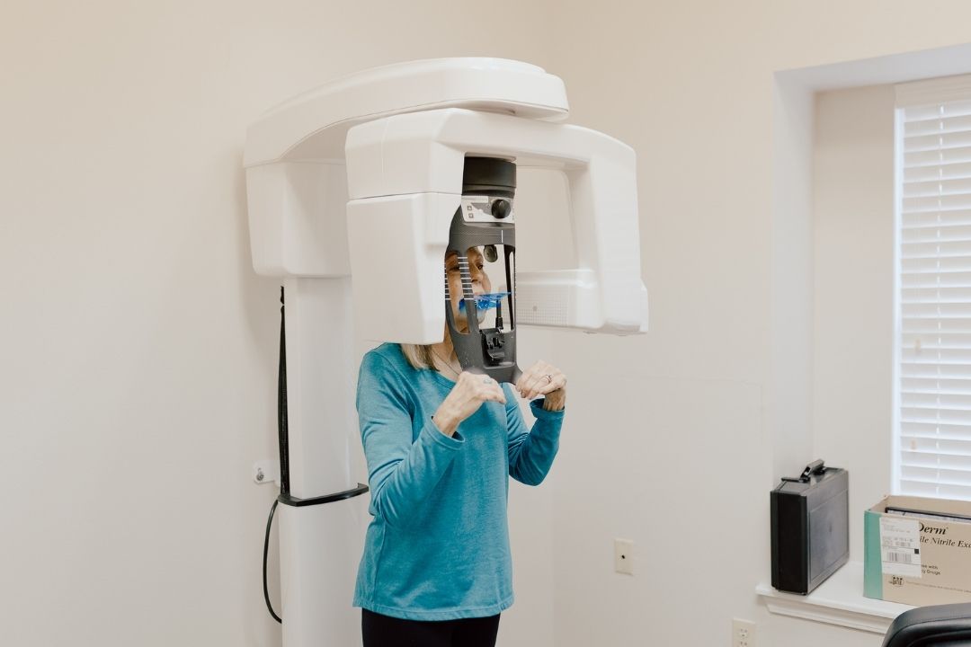 A person in a blue long-sleeve shirt is standing in a dental office, positioned in a panoramic dental X-ray machine, with their face partially obscured by the equipment.