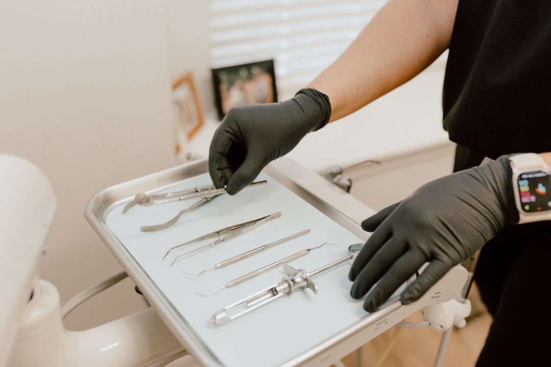 A person wearing black gloves arranges dental tools on a metal tray in a dental office. The tray holds forceps, tweezers, a mirror, and other dental instruments.