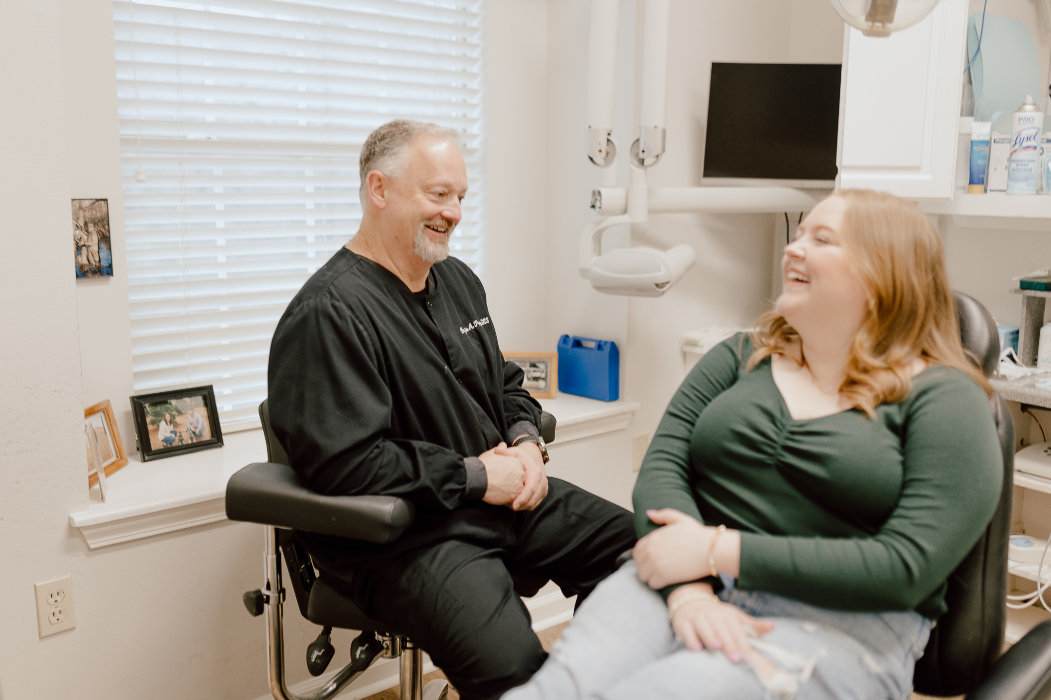 A dentist in black scrubs sits smiling and talking with a woman in a green top and jeans, who is laughing in the dental chair during her spring dental checkup. They appear relaxed in the bright, welcoming office.