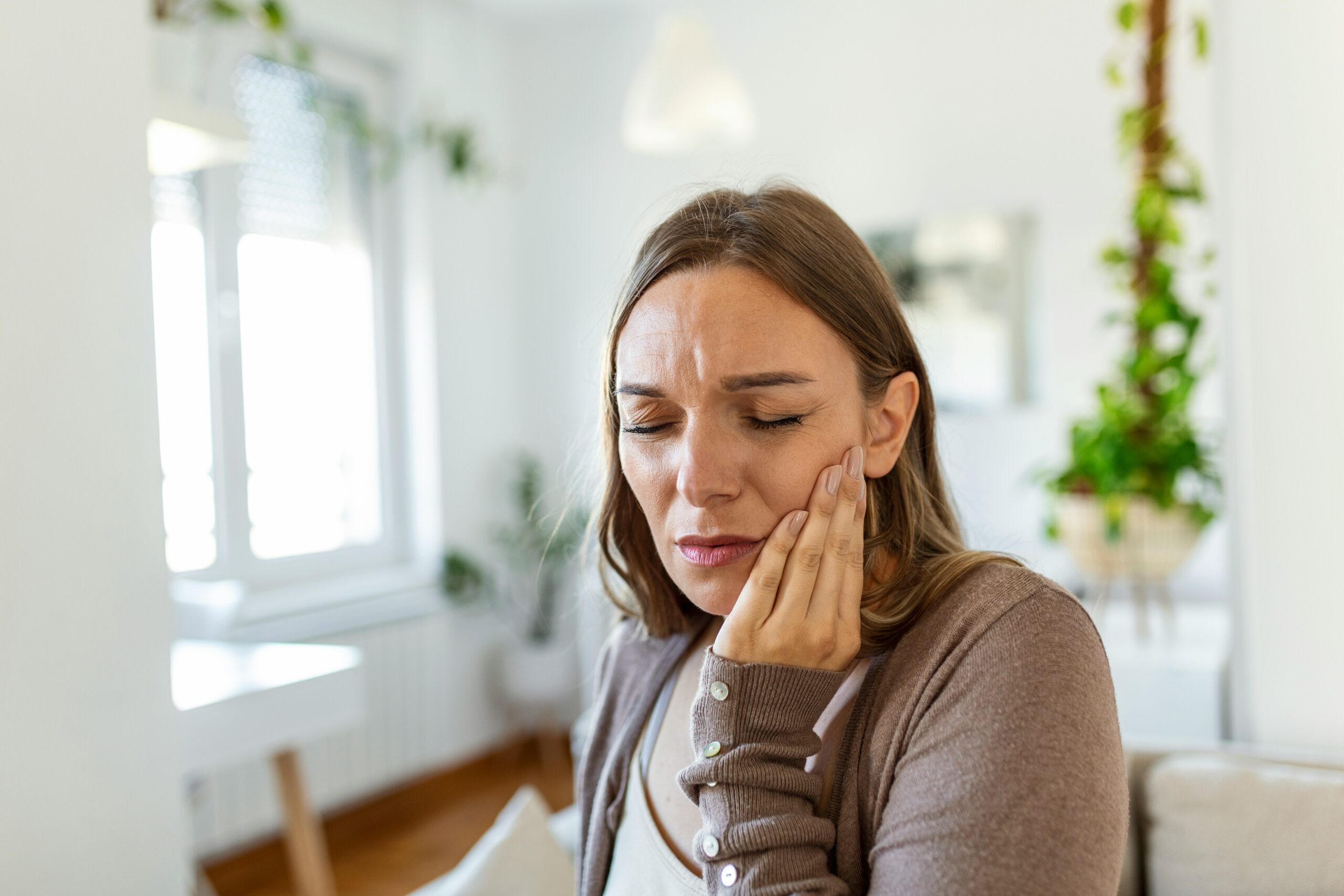 A woman sits indoors, holding her cheek with a pained expression that suggests she is suffering from a toothache. The background features bright, modern home decor with plants and large windows.