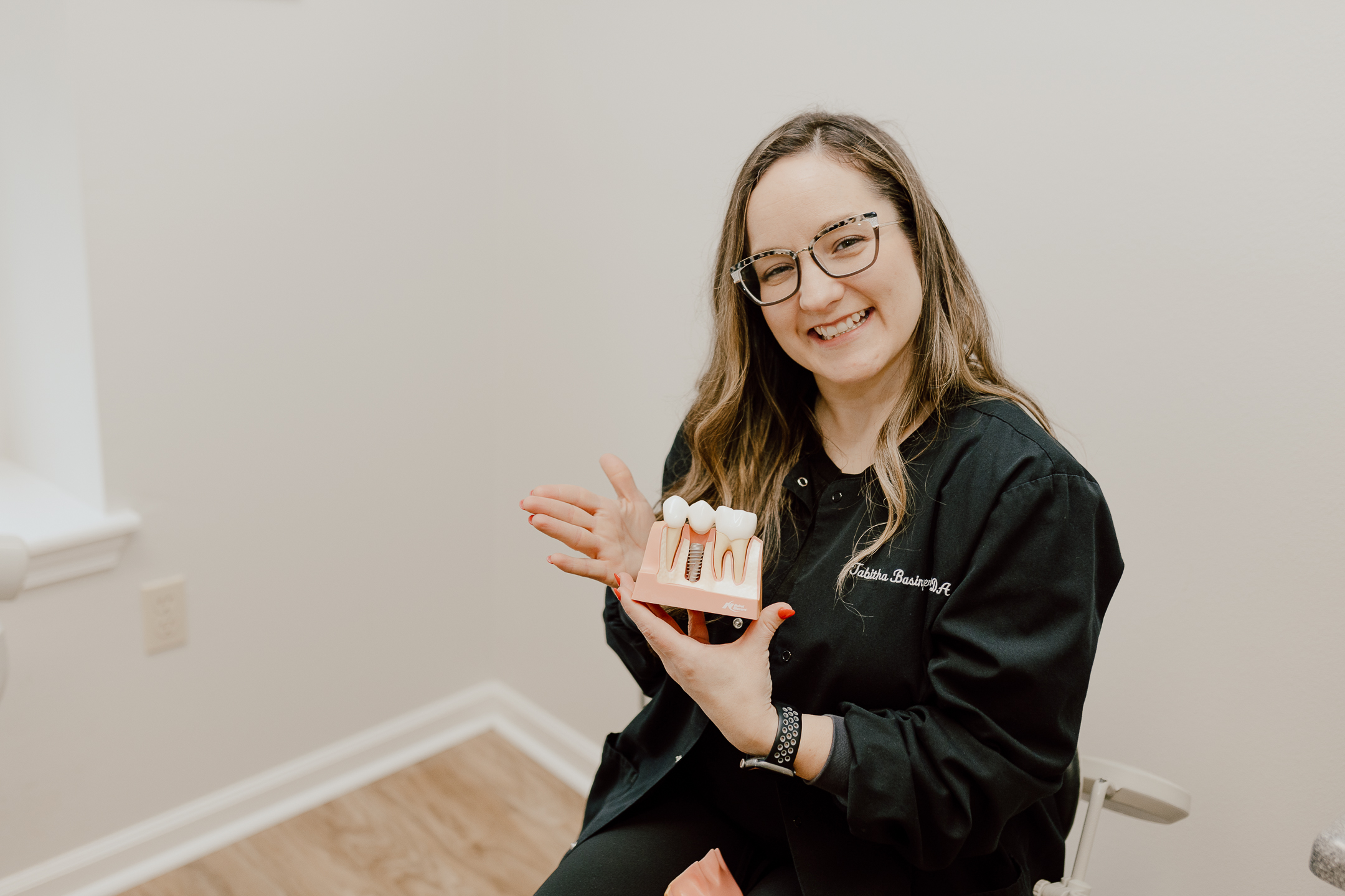 A smiling woman in glasses and a black scrub top holds a dental implant model, pointing to it with her left hand, showing what happens when there is delayed treatment with dental health