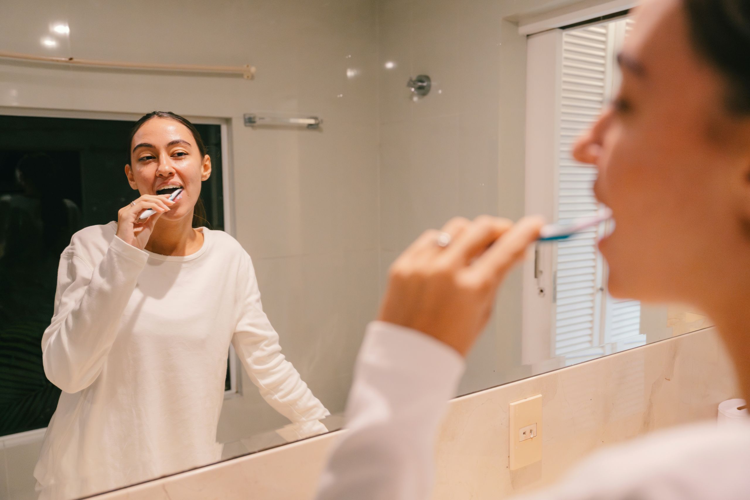 A person in a white shirt brushes their teeth while looking in the bathroom mirror. The reflection shows her smiling and also checking if can a tooth infection spread. The bathroom is well-lit with a window and white tile.