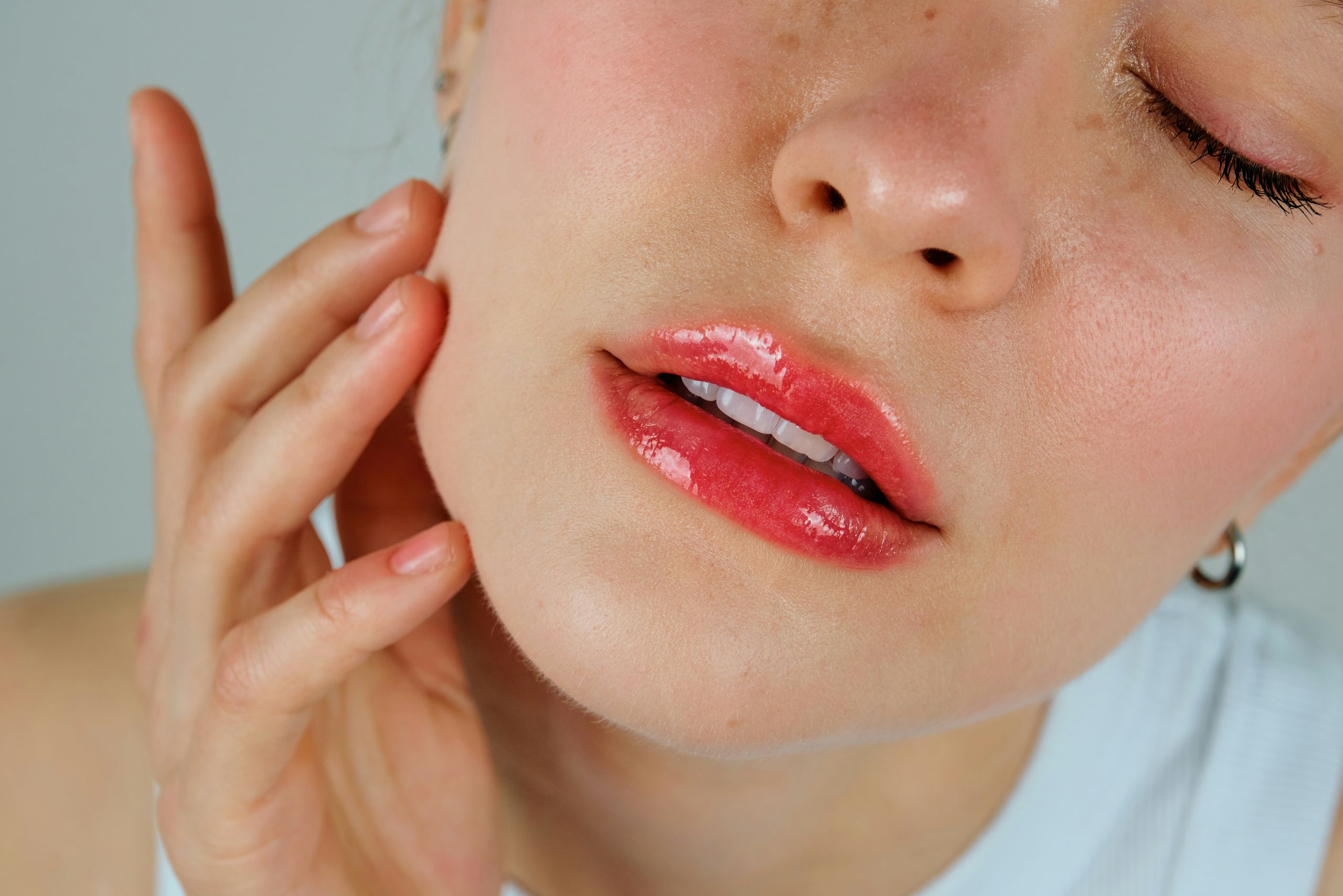 Close-up of a person with glossy pink lips, lightly touching their cheek with their fingers. Their eyes are closed and their facial expression appears to be troubled, like experiencing tooth pain when biting down
