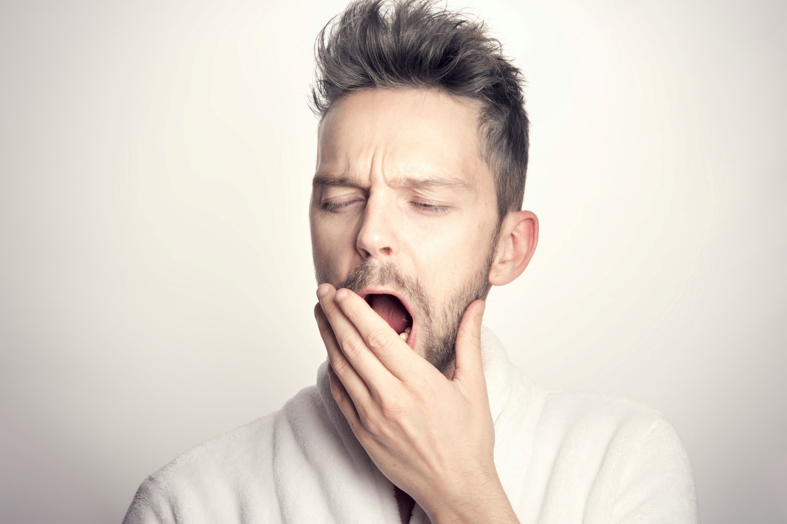 A man in a white robe yawns and covers his mouth with his hand, appearing tired or sleepy, against a plain light background,