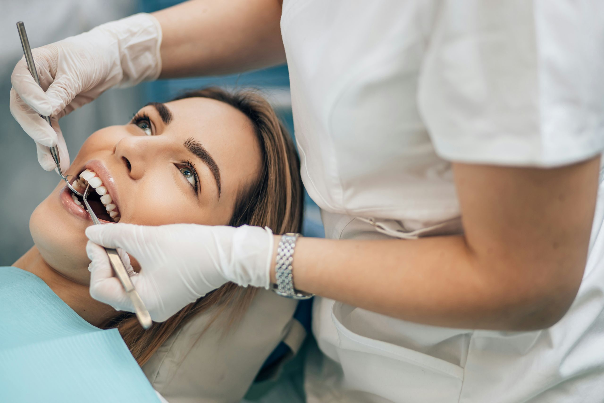 A dentist in white gloves examines a smiling womans teeth using dental tools while she lies in a dental chair.
