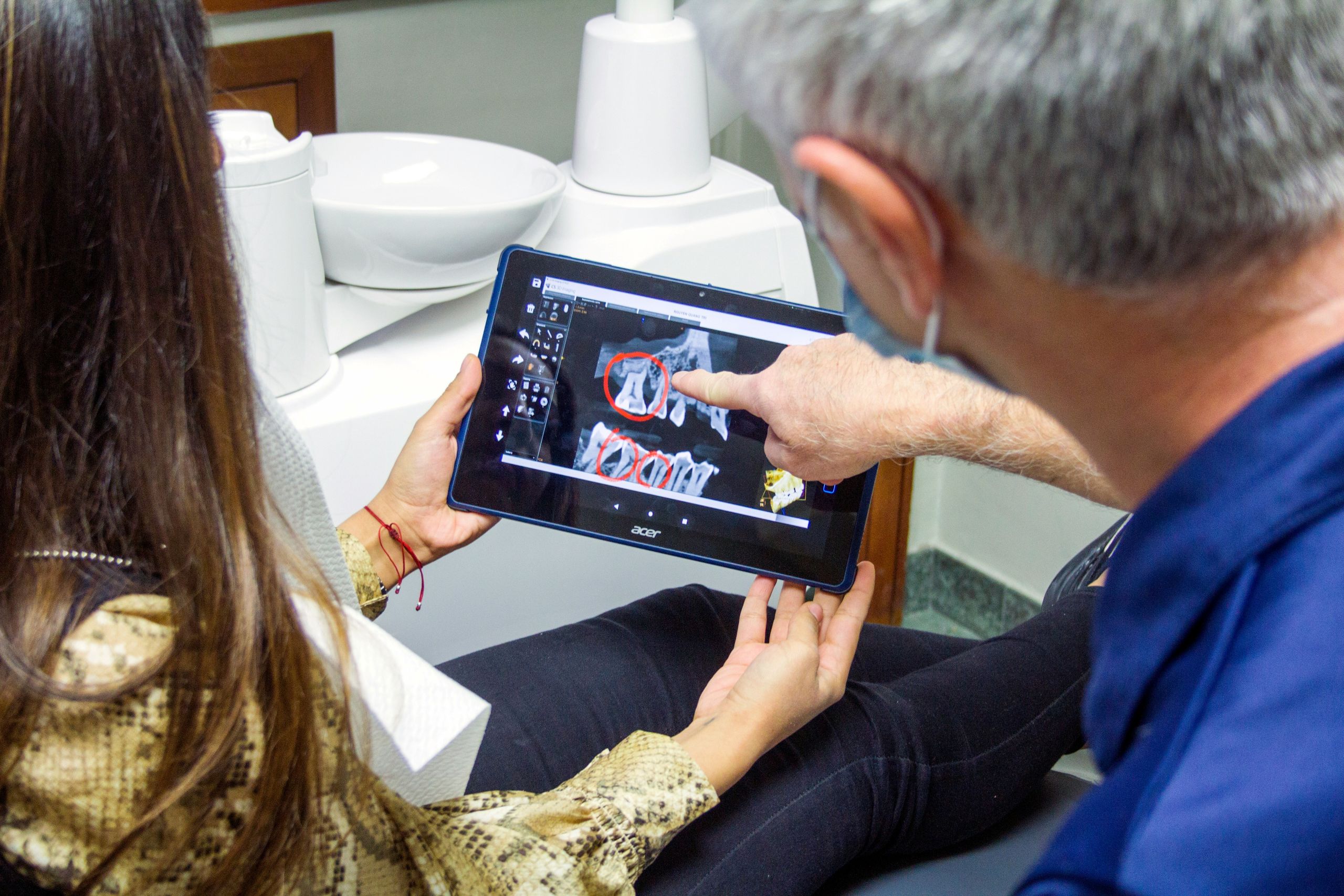 A dentist points to a digital X-ray of teeth on a tablet while consulting with a patient.
